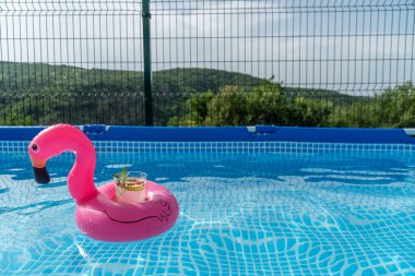 Şişirilebilir pembe flamingo havuz oyuncağının içindeki kokteyl, suyla dolu bir havuzda yüzüyor..