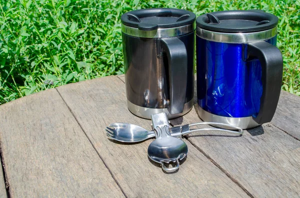Metal termo bardaklar, ahşap masada kaşıklar ve yeşil çimenler olan termo bardaklar.