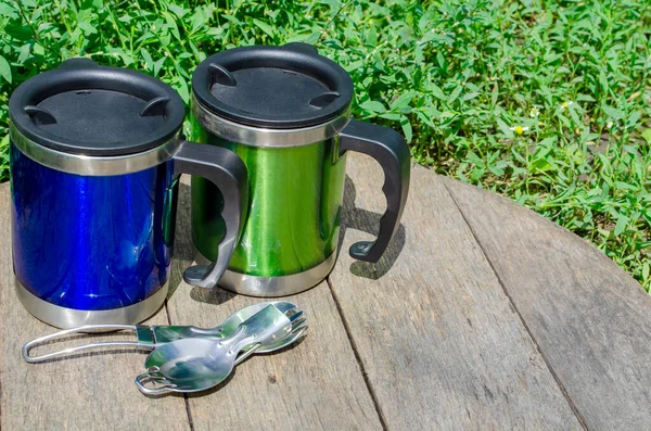 Metal termo bardaklar, ahşap masada kaşıklar ve yeşil çimenler olan termo bardaklar.