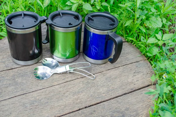 Metal termo bardaklar, ahşap masada kaşıklar ve yeşil çimenler olan termo bardaklar.