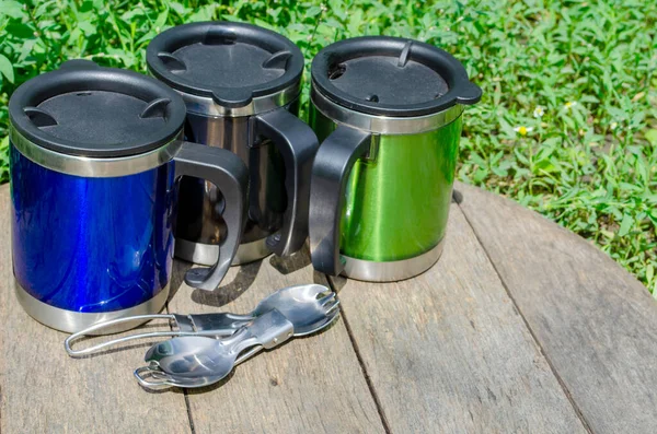 Metal termo bardaklar, ahşap masada kaşıklar ve yeşil çimenler olan termo bardaklar.