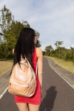 young girl with black hair walking on asphalt road, wearing red casual dress and backpack, tourist traveling and having fun on sunny day, lifestyle
