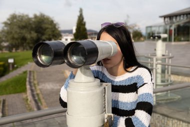 Uzun düz siyah saçlı çekici bir kadın. Parkta teleskop kullanıyor. Gündüz moda ve turist hayatı yaşıyor.