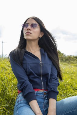 Genç bayan model parkta çimlerin üzerinde otururken rüzgar düz siyah saçlarıyla, güzellik ve doğa, yaşam tarzı ve moda