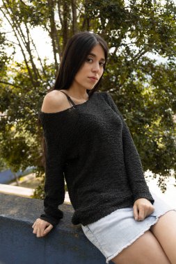 Doğayla iç içe bir parkta rahat bir şekilde poz veren kadın manken. Uzun siyah saçlı, rahat siyah etekli, hayat tarzı olan genç bir Latin kadın.