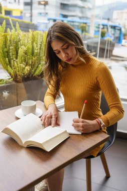 Sarı saçlı genç bir kız öğrencinin bir kitapla öğrendiği ve masaya bir fincan sıcak içecekle notlar aldığı bir gün. Yaşam tarzı, okuma ve hobi.