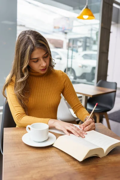 鉛筆で本を書きながら学び働く若いラテン語の美しい女性ブロンドの髪の長い女性コーヒーを飲みながらカフェテリアに座って学生生活 — ストック写真