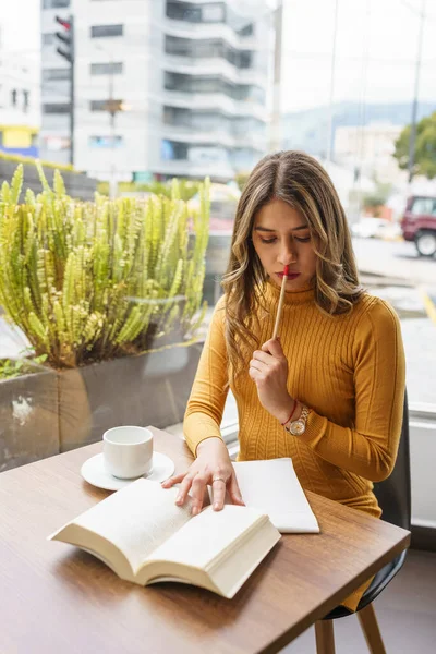 若いラテン語の女性で長いブロンドの髪を持つ職場は 集中するために熱い飲み物の横に ノートを作るためにペンで本に集中して座って — ストック写真