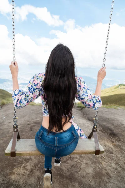 Salıncakta sallanan zayıf genç bir kadın, uzun düz saçları, günlük kıyafetleri, arka planı dağlarla ve bulutlarla kaplı gökyüzü, parkta yaşam tarzı ve eğlencesi var.