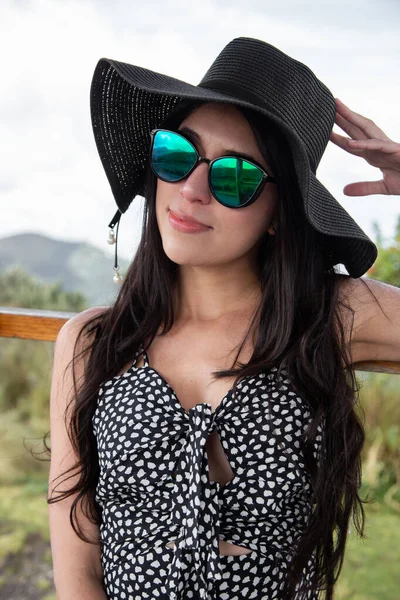Dağlı bir manzarada siyah saçlı, şapka, güneş gözlüğü ve uzun bir elbise giyen genç bir Latin kadın yaz için güzellik ve moda, yaşam tarzı ve turizm.