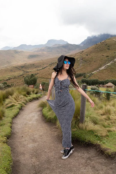 Doğanın ortasında yürüyen, uzun siyah saçlı, şapka takan, güneş gözlüğü takan, gündelik uzun elbiseli, gençlik güzelliği ve modası olan genç bir Latin kadın.