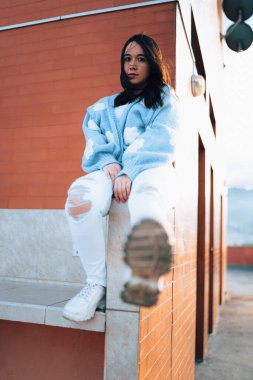 Ayakkabısının tabanını gösteren siyah saçlı ve doğal makyajlı genç bir Latin kadın, günlük kıyafetinde yün bir kazak giyiyor, gençlik modasında.