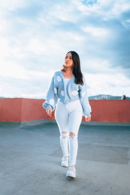 latin woman taking a walk outside, wearing a woolen jacket and casual jeans, background with sky and clouds, lifestyle and fashion trend
