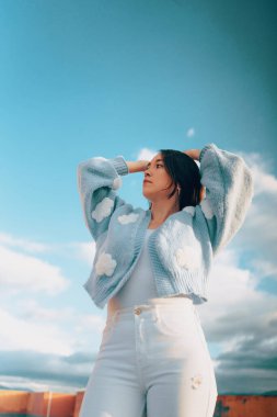 Model yaşam tarzı, modaya uygun yün süveter giyme, kollar havada poz verme, gökyüzü arka planı bulutlu, güzellik