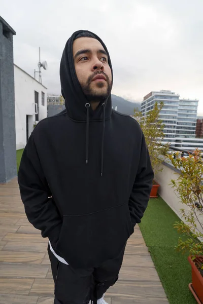 to protect himself from the cold he wears the hood of a sports sweater, urban fashion in the city a young latin brunette male model with a beard, lifestyle