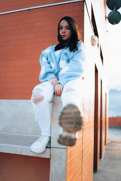 Ayakkabısının tabanını gösteren siyah saçlı ve doğal makyajlı genç bir Latin kadın, günlük kıyafetinde yün bir kazak giyiyor, gençlik modasında.