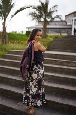 posing with her jacket a young latin brunette woman wears a long dress with a print skirt in a fashion, beauty and model lifestyle, in the background stands in the day