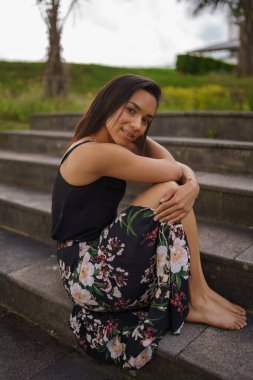 sitting hugging her legs a young brunette woman with short straight hair, wears a flower print skirt, summer fashion, lifestyle and beauty on the steps of a park