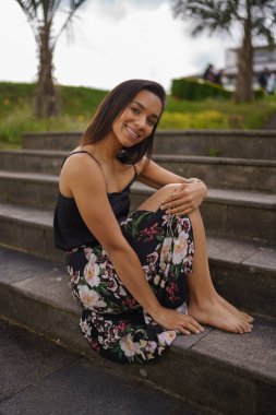 posing on a bleachers a young brunette woman with short straight hair, wears a flower print skirt, lifestyle, natural beauty and fashion