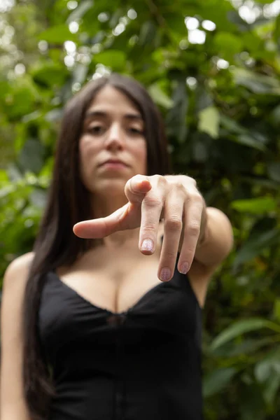 pointing to the camera with the index finger a young woman with straight black hair wears a black blouse with a neckline, nature and beauty of latin model in a park, lifestyle