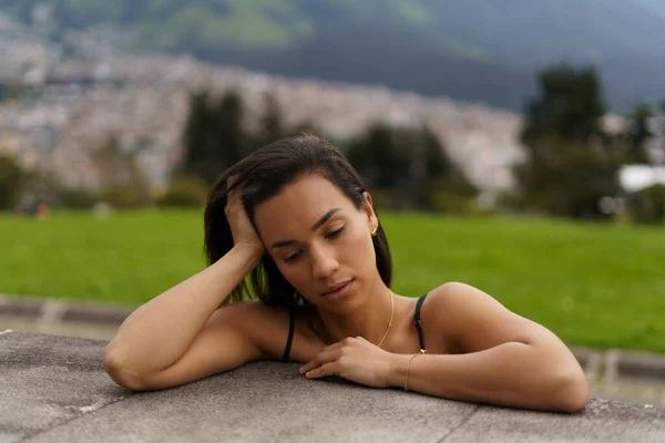 young latin brunette woman with short hair leaning against a sad platform, outside a park, mountain and city background, beauty