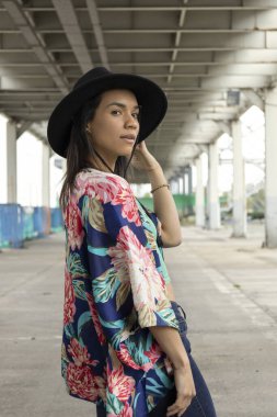 urban style of young latin brunette woman, natural beauty with fashionable accessories, model portrait, wearing a black hat and colorful kimono as an accessory