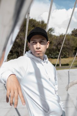 Relaxed by a Latin young boy using a casual cap and white hoddie, lifestyle and casual fashion in the city, sunny day in a park