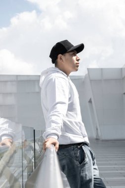 Standing with a profile face a young Latin man using a casual cap, white Hoddie jeans, casual lifestyle, natural beauty in the building background with crystals