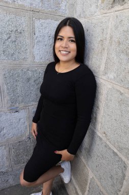 young latin woman with long black hair wearing casual black dress smiling and looking at camera, lifestyle and fashion, stone texture background, model beauty