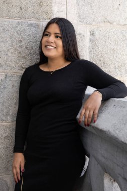 leaning against the wall a young latin woman with long black hair wearing a casual black dress smiling, lifestyle and fashion, stone texture background, model beauty