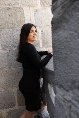 smiling and happy young latin woman with long black hair wearing a black dress, lifestyle and fashion, stone texture background, beauty
