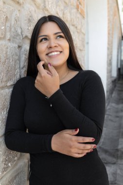 leaning against a stone wall, young slim woman with long hair wears a black dress, beauty with natural makeup in the daytime, joy