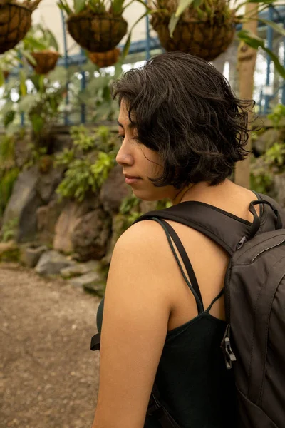 female traveler using a suitcase walking on a garden path surrounded by nature, young latin woman with short hair showing her face in profile, tourist lifestyle