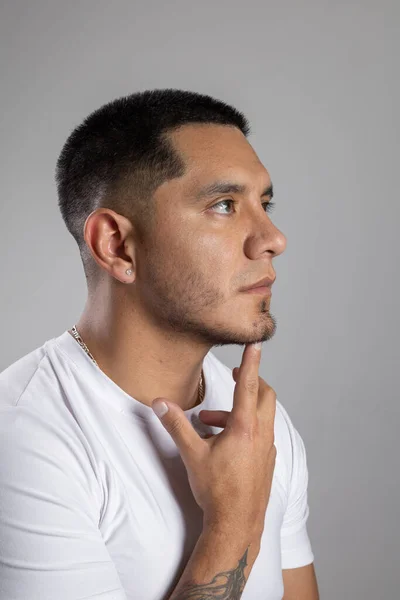 face of a young latin man with short urban style hair with a beard and a white t-shirt in studio has his index finger on his chin, thinking