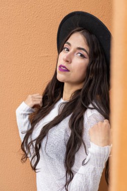 Stüdyo, turuncu arka planda, uzun dalgalı saçlı, aksesuar olarak şapka takan, Latin kadın modası ve güzelliği olan genç bir model.