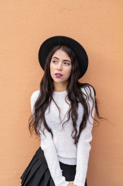 Stüdyoda turuncu arka planda genç bir kız şık bir şapka ve elbise giyiyor uzun dalgalı saçlı, güzel, manken bir Latin.