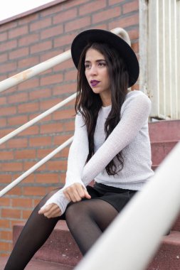 Açık tribünde dinlenen, şapka, yaşam tarzı ve güzelliği olan göz alıcı elbiseler giyen genç Latin bir kadın.