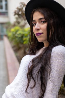 Güneşli bir günün tadını çıkaran uzun dalgalı saçlı, şapkalı, yaşam tarzı ve modası olan genç Latin bir kadın.