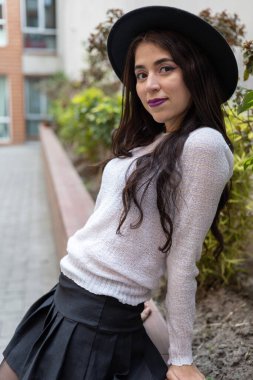 Kameraya bakıyor ve uzun dalgalı saçlı, rahat bir etek, süveter ve şapka giyen, gündüzleri gençlik yaşam tarzı ve moda ile gülümseyen Latin bir kadın.