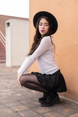 Gündüzleri şehvetli pozlar veren uzun dalgalı saçlı genç bir Latin kadın şapka, kazak, etek ve tayt giyer, moda ve güzellik, model yaşam tarzı