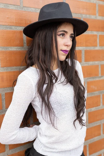 latin woman face with long hair, wear natural makeup and hat, brick wall background, beauty and accessory