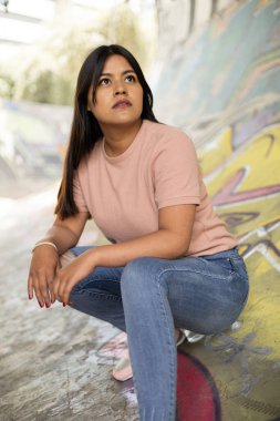 Doğal makyajlı, uzun düz saçlı, çömelmiş, günlük tişört giyen, yaşam tarzı, duvar yazısıyla dolu arka planı olan genç bir kadın.