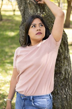 Bir ağaca yaslanmış, parkta güzel bir genç kadın, gündelik ve temel kıyafetlerle manken, gençlik yaşam tarzı.