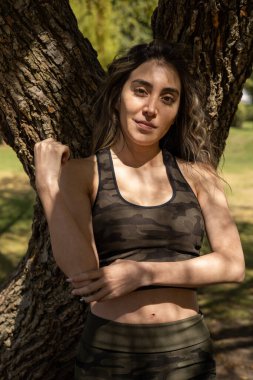 Bir ağacın gölgesinde duran kıvırcık saçlı, aktif spor giyimli, doğayla barışık genç bir kadın.