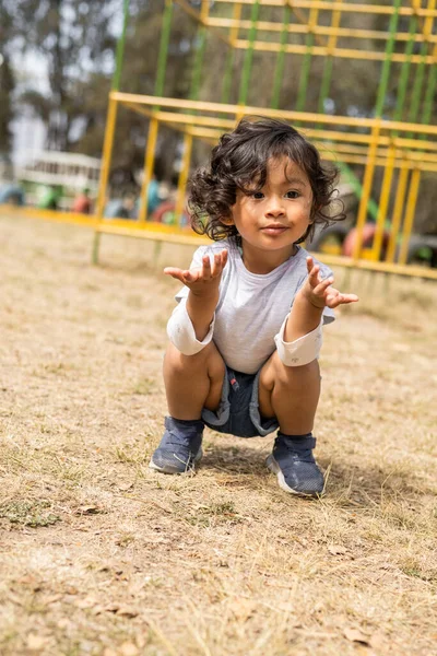 making gesture with hands while squatting beautiful little latin girl with curly hair, wear casual and summer clothes, childhood and lifestyle having fun in a park