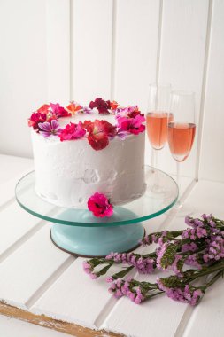 Beyaz kremalı ve yenilebilir çiçekli düğün pastasının güzel süslemesi, yanında iki kadeh şampanyalı, kutlamada tatlı olarak yiyecek, masa.