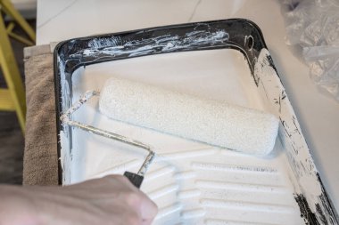 hand holds a paint roller in a paint tray with white paint for your project