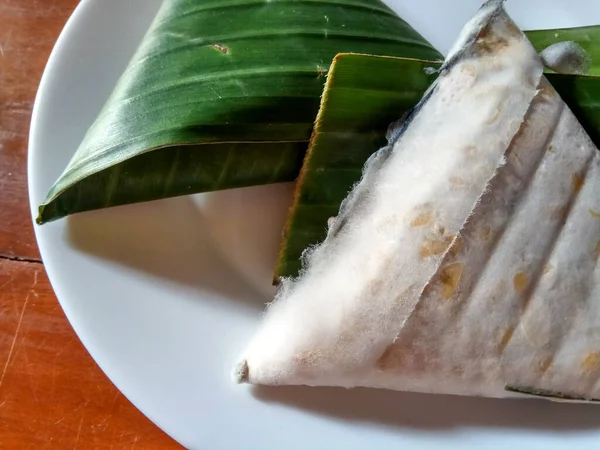 Tempeh or tempe, processed soy product, wrapped in banana leaves with triangular shape. Placed on white plate, with wooden background