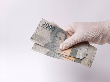 Beyaz lateks eldivenli bir elin içinde para, 2000 IDR, Endonezya banknotları var. Beyaz arka planda izole edilmiş. Yan görünüm.