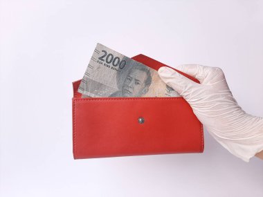 Beyaz lateks eldivenli bir elin içinde kırmızı bir çanta, içinde para, 2000 IDR ve Endonezya banknotları var. Beyaz arka planda izole edilmiş. Yan görünüm.
